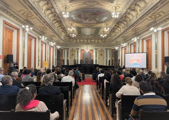 Auditório nobre com teto ornamentado e afrescos, paredes com molduras e luminárias pendentes. O espaço está cheio de pessoas sentadas, voltadas para o palco ao fundo, onde ocorre uma palestra. À direita, há um telão com a imagem do jornalista Guga Chacra e os dizeres “Encontros Acadêmicos”. O ambiente é solene e bem iluminado.