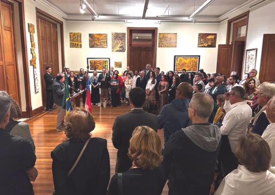 Durante o evento na galeria, o orador discursa ao lado das bandeiras do Brasil e do Chile, diante de um grande público atento. O salão tem piso de madeira, paredes com obras de arte e iluminação quente. As pessoas estão elegantemente vestidas, formando um semicírculo em torno do orador. O ambiente transmite formalidade e celebração cultural.