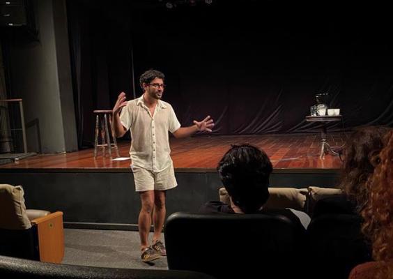 O ator Ari Areia se apresenta em frente ao palco do teatro, vestindo camisa e bermuda claras. Ele gesticula enquanto fala ao público, que o assiste de poltronas acolchoadas. O cenário é simples, com cortina preta ao fundo, um banco e uma mesa lateral. A iluminação foca no ator, criando uma atmosfera íntima e envolvente.