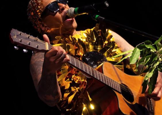 O cantor Àrt se apresenta no show Afetos Cítricos, usando um figurino dourado brilhante e óculos escuros. Ele canta ao microfone enquanto toca violão, com folhas verdes a frente do instrumento. A iluminação destaca sua performance intensa e expressiva contra o fundo escuro, criando uma atmosfera vibrante e performática.