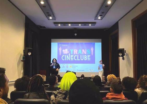 Público acompanha a abertura da 2ª edição do Trans Cineclube no Cinema. Duas pessoas falam ao microfone diante de uma tela que exibe o nome do evento e os logos dos apoiadores. A plateia, diversa e atenta, ocupa as poltronas acolchoadas. O ambiente é intimista, com iluminação suave e foco na projeção, destacando a importância do evento para a visibilidade e representatividade trans no audiovisual.