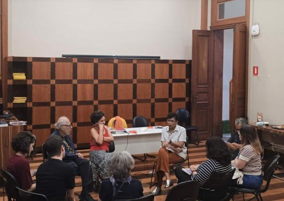Grupo participa de roda de conversa na Sala de Cursos com piso de madeira e prateleiras ao fundo. Duas pessoas conduzem o diálogo sentadas à frente, enquanto participantes escutam em semicírculo. O ambiente é acolhedor, com luz suave e mobiliário em madeira. Há materiais sobre a mesa, como livros e papéis.