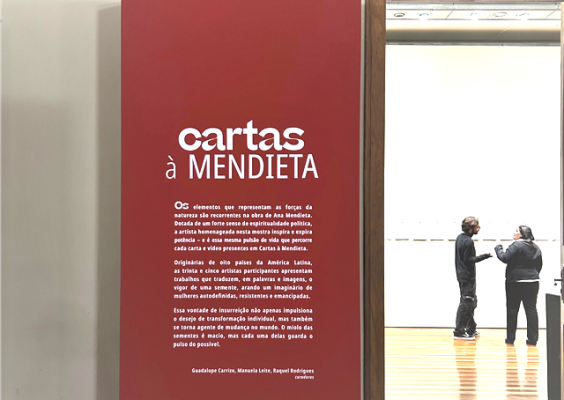 Totem vermelho exibe texto de apresentação da mostra “Cartas à Mendieta” na Galeria de Exposições com piso de madeira. Ao fundo, duas pessoas observam e conversam diante de obras expostas em paredes brancas. O ambiente tem iluminação clara, aspecto minimalista e acolhedor, destacando o caráter reflexivo e artístico da exposição, que homenageia Ana Mendieta reunindo cartas e vídeos de artistas latino-americanas.