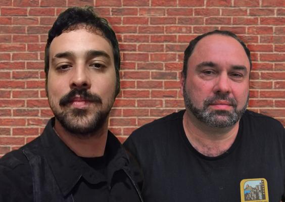 Guilherme Guimarães e Andre Angulo diante de fundo de parede de tijolos. Guilherme, à esquerda, tem barba e bigode curtos, cabelo escuro e veste camisa preta. Andre, à direita, tem barba cheia grisalha, cabelo curto escuro e veste camiseta preta com logo “Favela República” no peito. Ambos olham diretamente para a câmera, com expressões neutras.