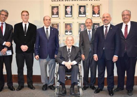 Oito homens posam para foto formal em frente à “Galeria dos Diretores do CCJF”. Sete estão de pé, vestindo ternos escuros; ao centro, um homem idoso está sentado em uma cadeira de rodas, também de terno. Atrás deles, há retratos emoldurados na parede com fotos de ex-diretores da instituição. O ambiente é interno, com iluminação clara e piso cinza. A atmosfera é solene e institucional.