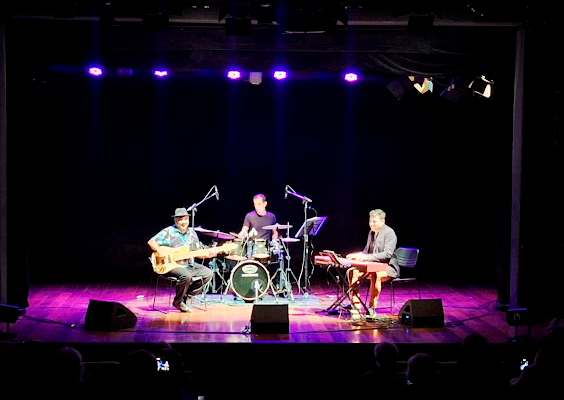 A imagem registra o show “50Tão”, do Ney Conceição Trio, no palco do Teatro do Centro Cultural Justiça Federal (CCJF). Sob luzes violetas, três músicos se apresentam: Ney Conceição no contrabaixo à esquerda, acompanhado por um baterista ao centro e um tecladista à direita. O fundo escuro destaca a iluminação do palco, refletida no piso de madeira. Na plateia, algumas pessoas assistem e registram o momento com celulares.