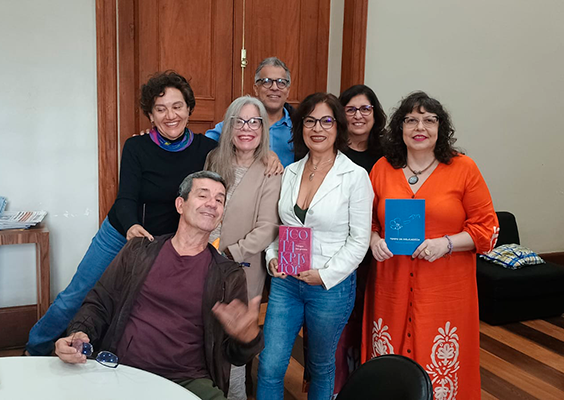 A imagem mostra sete pessoas reunidas em uma sala com piso de madeira e portas altas de madeira ao fundo, durante um encontro em homenagem à escritora suíça-húngara Agota Kristof. Seis delas estão em pé, sorrindo, enquanto uma posa de forma descontraída sentada à frente. Duas mulheres seguram livros — um deles, com o nome da autora em destaque. O clima é acolhedor e celebra a literatura e a memória da homenageada.
