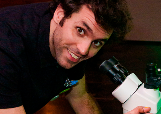A imagem mostra Daniel Diaz, Diretor, Coordenador Geral, Curador e Produtor do Festival ECRÃ. Ele aparece sorridente, com expressão simpática, enquanto manuseia um microscópio branco em um ambiente com iluminação colorida. A cena sugere uma atmosfera descontraída e criativa, alinhada ao espírito inovador do festival que promove a interseção entre arte e tecnologia.