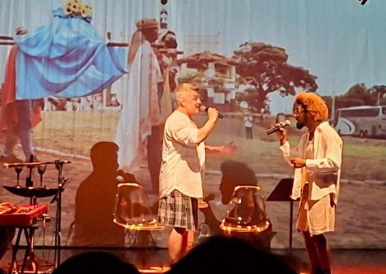 Fotografia do show do Trio Minas Portugal no CCJF. À esquerda, Francisco Eiras toca teclado eletrônico, enquanto, ao centro, Alessandro Brandão e, à direita, Cleiton Sobreira cantam, cada um segurando um microfone. Ao fundo, é projetada uma imagem colorida de cena ao ar livre, com figuras fantasiadas, compondo o cenário artístico da apresentação.