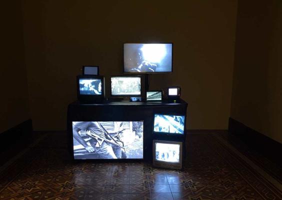Conjunto de televisores e monitores de diferentes tamanhos exibindo imagens em preto e branco e coloridas, dispostos sobre uma estrutura escura em uma sala de piso decorado. A instalação é Nine Lives Cat, apresentada na Galeria da Cela do CCJF durante o Festival ECRÃ.