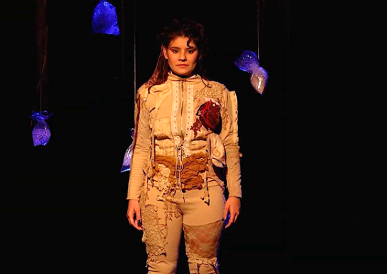 A imagem mostra uma cena da peça teatral Na Quinta Dor, apresentada no Teatro do Centro Cultural Justiça Federal (CCJF) durante o mês de julho. No centro do palco, uma atriz está sob iluminação pontual, vestindo um figurino claro com texturas e bordados artesanais. Um coração em relevo se destaca no lado esquerdo do peito. Ao fundo, sacos plásticos translúcidos pendem do teto, iluminados em azul, criando uma atmosfera simbólica e impactante.