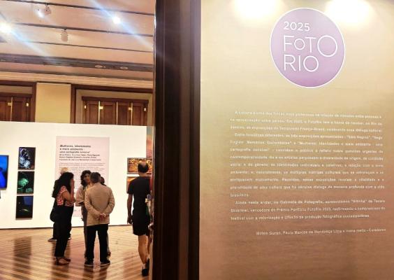 A imagem mostra uma exposição de fotografia chamada "FotoRio 2025", realizada em um espaço interno com piso de madeira e iluminação de teto. Visitantes observam painéis com fotos coloridas e textos explicativos. Na parede à direita, há um painel com o logotipo do evento e um texto introdutório sobre as temáticas abordadas na exposição, como cultura, identidade, gênero e meio ambiente.