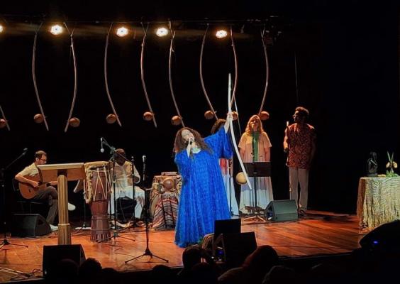 Em um palco iluminado, uma cantora de vestido azul longo se apresenta segurando um berimbau. Atrás dela, há músicos tocando violão, percussão e duas pessoas em pé próximas a microfones, como coristas. No fundo, vários berimbaus estão pendurados verticalmente. À direita, uma mesa coberta por tecido com flores e um objeto decorativo. O público aparece em silhueta na parte inferior da imagem.