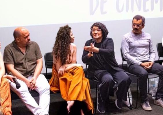Seis pessoas estão sentadas lado a lado em cadeiras no palco de um auditório. Elas participam de uma conversa em grupo, com microfone em mãos, diante de uma tela de projeção. Uma mulher de roupa preta fala enquanto os demais escutam atentamente. A cena sugere um debate ou mesa-redonda ligada ao cinema ou à cultura.
