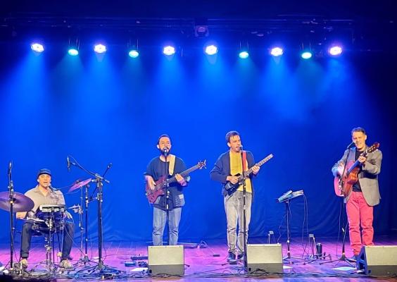 Quatro músicos se apresentam em um palco iluminado com luzes azuis e roxas. À esquerda, um baterista toca enquanto três instrumentistas cantam ao microfone: um com contrabaixo, outro com cavaquinho e o terceiro com violão. Todos estão posicionados em linha à frente de microfones, transmitindo a atmosfera de um show ao vivo.