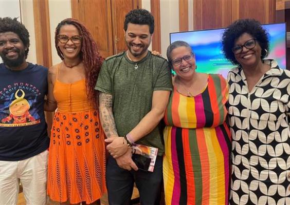 Cinco pessoas estão lado a lado, sorrindo para a foto em ambiente interno. À esquerda, um homem de camiseta azul com estampa colorida, seguido por uma mulher de vestido laranja, um homem de camiseta verde, uma mulher de vestido listrado em cores vibrantes e outra de vestido preto e branco estampado. Todos parecem descontraídos e felizes, transmitindo clima de amizade e celebração.
