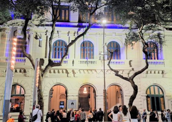 Fachada iluminada do Centro Cultural Justiça Federal à noite. O prédio histórico, em tom claro, exibe janelas arqueadas e detalhes clássicos, realçados por luzes azuladas. Árvores altas moldam a cena. Sob os três arcos de entrada, um grande lustre é visível no interior. Na calçada, muitas pessoas circulam e conversam, criando clima de evento.