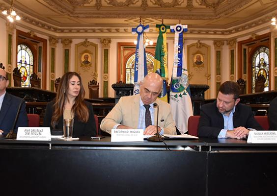 Cinco pessoas estão sentadas lado a lado em uma mesa de cerimônia em um salão histórico com vitrais e bandeiras ao fundo. No centro, o diretor-geral do CCJF, Theophilo Miguel, assina um documento. À sua direita estão a desembargadora estadual Ana Cristina Dib Miguel e outro homem; à esquerda, Nicola Miccione, secretário de Estado da Casa Civil, e outro participante. Todos usam trajes formais.