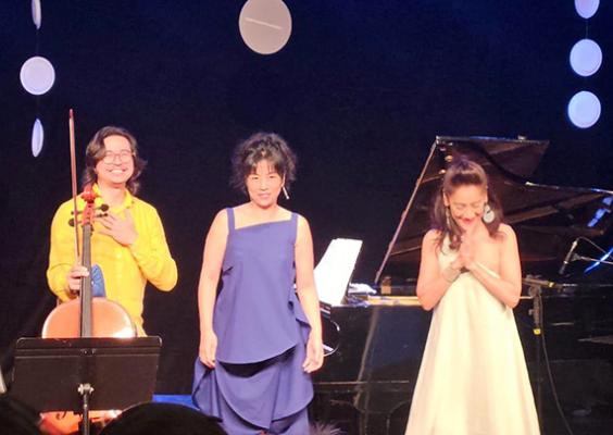 Três musicistas se apresentam no palco durante o concerto Brasil Japão – Atravessando o Tempo e o Oceano. À esquerda, um deles segura um violoncelo; ao centro e à direita, as outras duas artistas se posicionam próximas a um piano de cauda. As três se curvam em agradecimento ao público, sob iluminação suave e cenário com elementos circulares brancos suspensos, que reforçam o clima de elegância e harmonia do espetáculo.