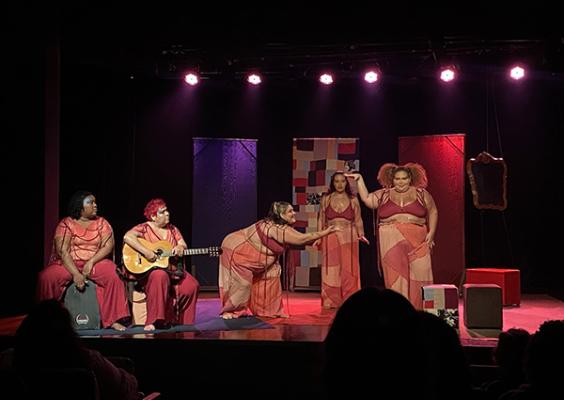 Cena da peça teatral Deixa eu dizer que te amo, com cinco mulheres negras no palco, vestidas com figurinos em tons de rosa e vermelho. Uma das artistas toca violão enquanto as demais interagem de forma expressiva e afetuosa. O cenário apresenta painéis coloridos e um espelho lateral, sob iluminação rosada que cria um clima acolhedor e poético. A plateia acompanha atenta a encenação.