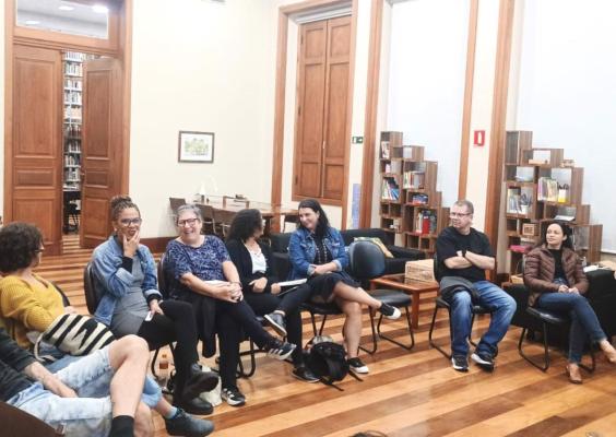 Homens e mulheres sentados em cadeiras pretas (posicionadas no formato de meia lua) conversam em uma sala com piso de madeira e grandes portas de madeira.