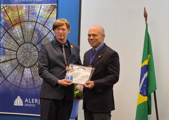 Dois homens brancos, de terno, sorriem para as câmeras com um certificado em mãos. Do lado direito, a bandeira do Brasil; no lado esquerdo, um cartaz com uma imagem semelhante a uma rosácea, cinza.
