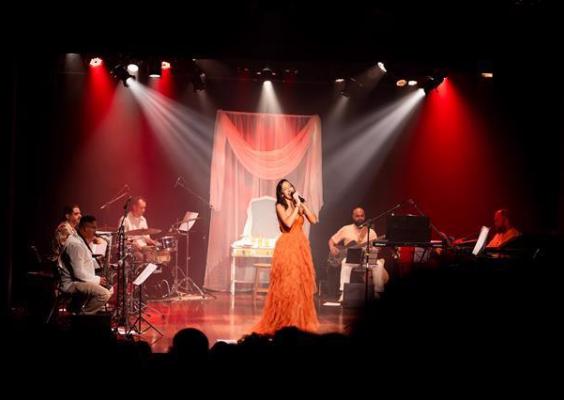Cantora de vestido laranja se apresenta no palco iluminado por luzes vermelhas e brancas, acompanhada por banda com teclado, baixo, bateria e saxofone diante de plateia.