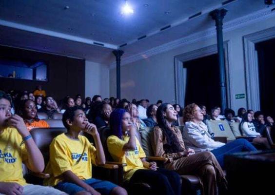 Público em auditório assiste atento a uma exibição. Nas primeiras fileiras, crianças com camisetas amarelas “Super Hacka Kid”; atrás, adultos e jovens observam a tela iluminada por luz azul.