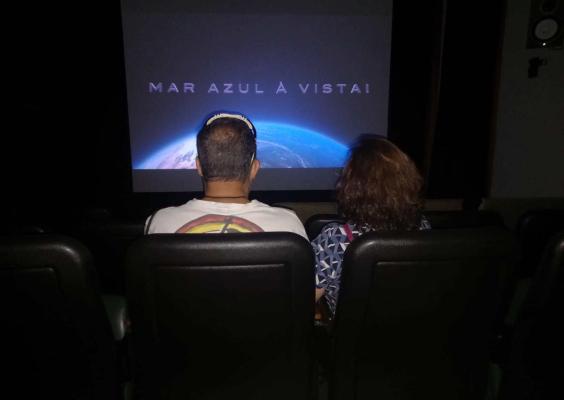 Homem e mulher estão de costas, sentados em poltronas pretas acolchoadas em uma sala de cinema. Na tela, à frente deles, uma cena de parte do planeta Terra com a frase "Mar Azul à Vista".