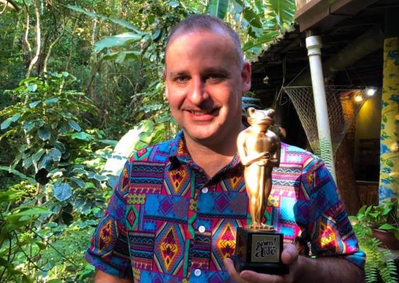 Alexandre Juruena, diretor-geral e curador do festival Anim!Arte, sorri segurando um troféu dourado com formato que remete a uma figura egípcia. Ele veste uma camisa colorida de estampas geométricas e está em um ambiente externo cercado por vegetação tropical, sob luz natural.