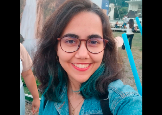 Fotografia da entrevistada Luise França sorrindo, usando óculos e jaqueta jeans, em ambiente externo.