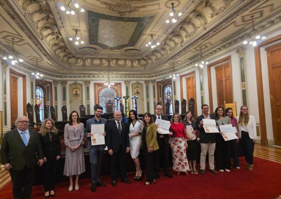 Foto em um grande salão histórico, com teto alto e ornamentado, lustres acesos, vitrais e cadeiras de madeira ao fundo. Um grupo de cerca de 14 pessoas, mulheres e homens adultos, está alinhado sobre um tapete vermelho, alguns segurando certificados. Eles posam sorrindo para a câmera, em clima de cerimônia ou premiação oficial.