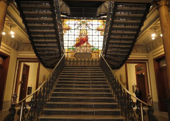 Escadaria monumental do CCJF vista de frente, com degraus largos que se dividem em duas rampas laterais ornamentadas por corrimãos trabalhados em metal. Ao fundo, um grande vitral colorido ilumina o ambiente com luz quente, destacando figuras e tons vibrantes. A cena transmite imponência e caráter histórico.