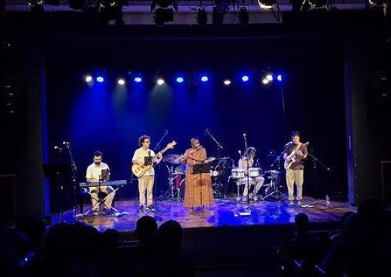 A imagem mostra um grupo de seis músicos se apresentando em um palco iluminado por luzes azuis. Da esquerda para a direita, há um tecladista, um guitarrista, uma flautista ao centro, seguida por um baterista, um percussionista e um saxofonista. Eles tocam diante de uma plateia em um teatro, com microfones e equipamentos distribuídos pelo palco. A cena transmite clima de show ao vivo e envolvimento musical.