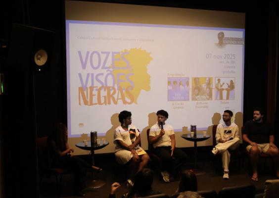 Fotografia do cinebate Vozes e Visões Negras no teatro do CCJF. A imagem mostra quatro participantes sentados no palco, conversando diante de uma tela de projeção que exibe o nome do evento e imagens relacionadas. Parte da plateia aparece em primeiro plano, acompanhando a discussão sobre protagonismo negro no audiovisual.