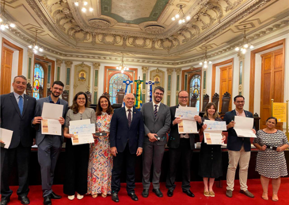 Na Sala de Sessões do CCJF, um grupo de finalistas, autoridades e jurados posa para a foto oficial da cerimônia do concurso Quis Ego Sum. Todos estão alinhados ao centro da sala ornamentada, sob um teto ricamente decorado e entre paredes clássicas. Alguns participantes exibem certificados enquanto sorriem, sobre um piso de madeira com um grande tapete vermelho.