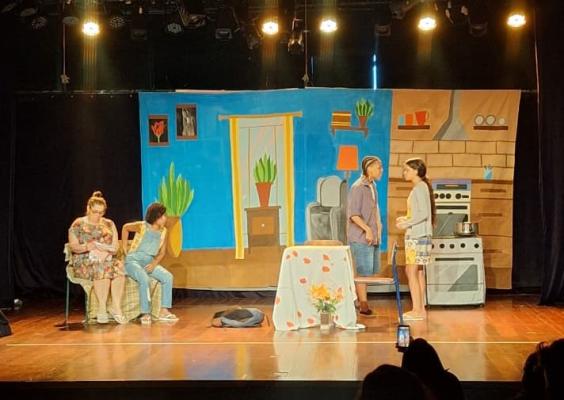 Palco de teatro com cenário colorido que representa o interior de uma casa. Ao fundo, uma parede pintada com porta, quadros, plantas, sala e cozinha com fogão e armários. À esquerda, duas pessoas sentadas em cadeiras: uma mulher adulta e uma jovem, próximas e em atitude de escuta. Ao centro, uma mesa pequena coberta com toalha clara e um vaso de flores. À direita, duas mulheres em pé conversam frente a frente, perto do fogão. A iluminação é quente e o público aparece em sombra na plateia.