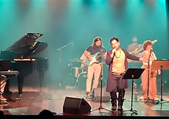 A imagem registra uma apresentação musical em palco, com um grupo de artistas distribuídos entre instrumentos e microfones. Ao centro, o cantor se posiciona à frente, interpretando a canção, enquanto músicos o acompanham ao piano, guitarra, baixo, bateria e vocais de apoio. A iluminação suave e a disposição equilibrada dos performers criam um ambiente intimista, destacando a interação entre os músicos e a atmosfera cênica do espetáculo.