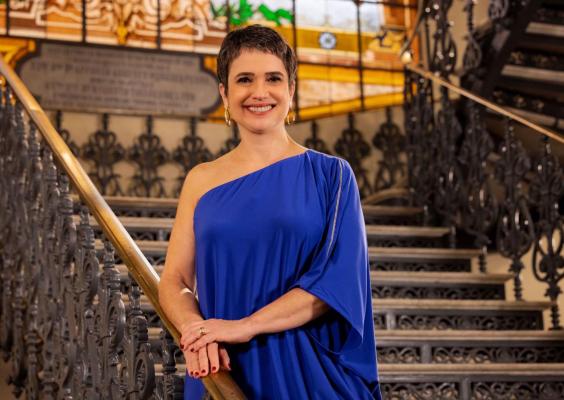 A imagem mostra a jornalista Sandra Annenberg posicionada ao centro da escadaria principal do CCJF, apoiando as mãos no corrimão de metal ornamentado. Ela veste um traje azul de um ombro só e sorri para a câmera. Ao fundo, destacam-se os degraus amplos, as grades trabalhadas e os elementos arquitetônicos clássicos do edifício histórico, compondo um ambiente solene e institucional.