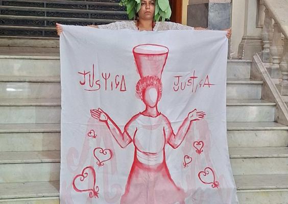 A imagem mostra uma mulher posicionada no centro de uma escadaria de mármore, segurando um grande tecido branco ilustrado com uma figura simbólica em tons de vermelho e a palavra “Justiça” escrita nas laterais. Ela veste roupa clara e traz folhas verdes sobre a cabeça, compondo uma referência ritualística. Ao fundo, destacam-se os degraus, o corrimão e os elementos arquitetônicos clássicos, que reforçam o caráter solene e simbólico da cena.
