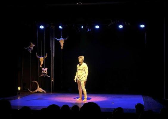 A imagem registra uma cena da peça A Hora do Boi, apresentada no CCJF. No centro do palco, o ator se posiciona sob iluminação cênica, destacando sua presença corporal e expressão dramática. Ao redor, elementos cenográficos suspensos, como formas que remetem a chifres e figuras simbólicas, compõem um ambiente ritualístico. A atmosfera intimista e a relação direta com a plateia reforçam o caráter simbólico e performático do espetáculo.