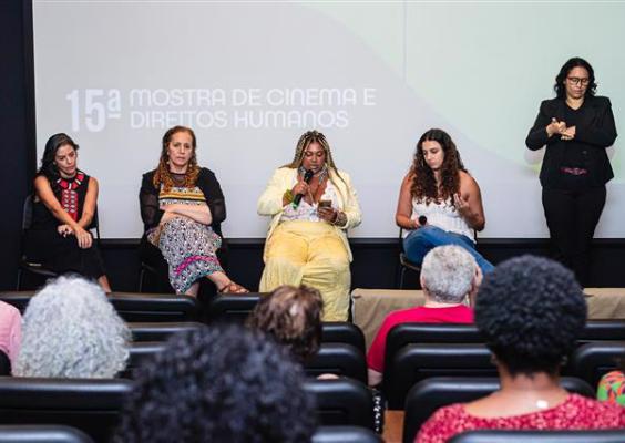 A imagem registra uma mesa de debate da 15ª Mostra Cinema e Direitos Humanos, com cinco mulheres sentadas à frente do público, em uma sala de cinema. Ao fundo, a projeção identifica o evento, enquanto uma intérprete de Libras acompanha a discussão. As participantes seguram microfones e papéis, em postura atenta e reflexiva. A composição evidencia o caráter formativo do encontro e o diálogo direto com a plateia presente.
