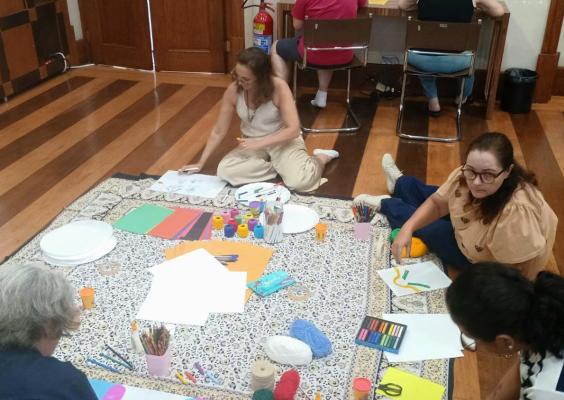 A imagem mostra um grupo de mulheres reunidas em uma atividade artística coletiva, sentadas no chão ao redor de um grande tecido que serve de apoio para os materiais. Papéis coloridos, tintas, lápis, pincéis e linhas estão espalhados, compondo um ambiente de criação e troca. O espaço interno, com piso de madeira e mesas ao fundo, reforça o caráter acolhedor e formativo da oficina, marcada pela interação e pela expressão criativa.