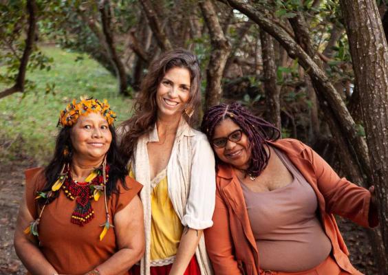 A imagem mostra três mulheres em ambiente natural, posicionadas lado a lado entre árvores e vegetação. Elas sorriem para a câmera, transmitindo proximidade e acolhimento. A mulher à esquerda usa adornos coloridos e um cocar floral, a do centro veste roupas claras com tons amarelos, e a da direita usa óculos e roupas em tons terrosos. O cenário arborizado e a luz natural criam uma atmosfera leve, afetiva e integrada à natureza.