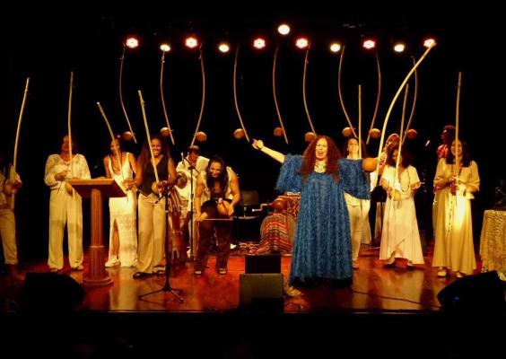 A imagem mostra um grupo de artistas reunidos no palco, sob iluminação quente, durante uma apresentação coletiva. Ao centro, uma mulher de vestido azul ergue o braço em gesto expressivo, enquanto os demais participantes, vestidos em tons claros, seguram berimbaus e instrumentos de percussão. A disposição cênica, a presença dos instrumentos e a interação entre os corpos reforçam o caráter ritualístico e celebrativo da performance.