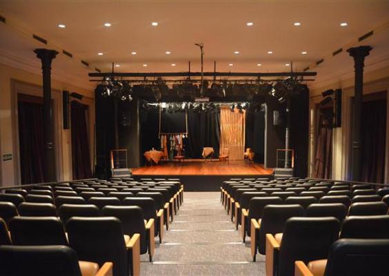 A imagem mostra o teatro do CCJF visto a partir da plateia, com fileiras de poltronas alinhadas em direção ao palco. A iluminação cênica destaca o espaço de apresentação, onde se veem elementos de cenário e equipamentos técnicos. As colunas laterais, o acabamento clássico e a organização do ambiente reforçam o caráter institucional e acolhedor do teatro, preparado para receber espetáculos, apresentações musicais e atividades culturais.