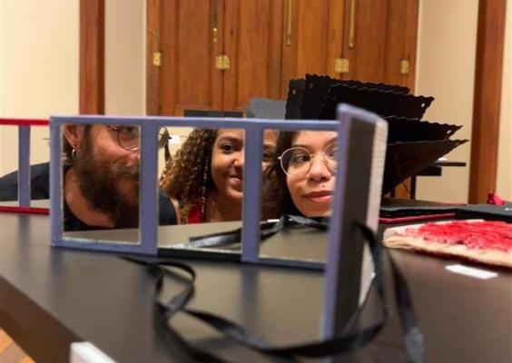 A imagem mostra três pessoas posicionadas atrás de uma mesa expositiva, com seus rostos refletidos em pequenos espelhos retangulares dispostos em sequência. Os reflexos fragmentados criam um jogo visual que destaca expressões atentas e curiosas. Em primeiro plano, a estrutura dos espelhos organiza a cena, enquanto, ao fundo, as portas de madeira e os detalhes arquitetônicos clássicos situam a instalação em um ambiente institucional e histórico.