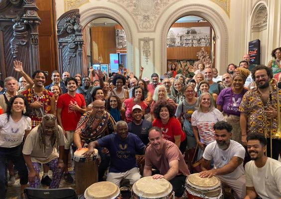 A imagem registra o público e os integrantes da Orquestra Balogun reunidos após a apresentação, ocupando o saguão de um edifício histórico. Músicos e espectadores posam juntos, sorridentes, com tambores e instrumentos de percussão em primeiro plano. Ao fundo, os arcos ornamentados, esculturas e detalhes arquitetônicos clássicos reforçam o caráter institucional do espaço, enquanto a diversidade de pessoas evidencia o clima de celebração, encontro e partilha promovido pelo espetáculo.
