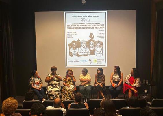 Roda de conversa “Vozes pelo fim do feminicídio e outras violências contra a mulher”, no cinema do CCJF. Sete mulheres estão sentadas no palco, em frente a uma plateia, participando do debate. Ao fundo, uma tela exibe o cartaz do evento. O ambiente é escuro, com iluminação focada nas participantes, criando clima de escuta e reflexão.