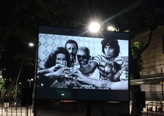 Tela de cinema ao ar livre instalada em uma rua arborizada durante a noite. Na projeção em preto e branco, um grupo de pessoas brinda com copos erguidos, reunidas em clima festivo. Luzes fortes iluminam a estrutura da tela, enquanto árvores, grades e caixas de som aparecem ao redor. Ao fundo, a rua escura e silenciosa reforça o clima cultural e urbano da cena.